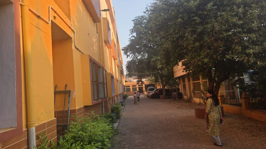 A brick path between yellow buildings and trees inside the Mumukshu Bhawan campus, Varanasi